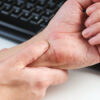 A man massaging his painful wrist against a background of a computer keyboard, illustrating the discomfort of carpal tunnel syndrome. Close up.