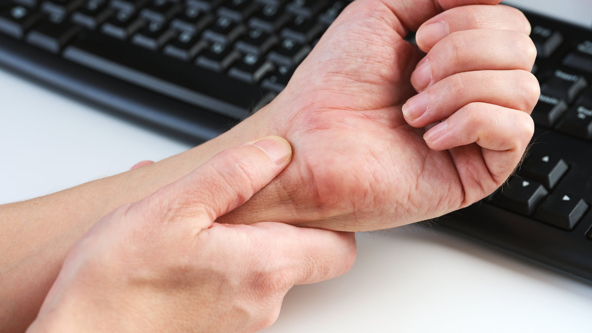 A man massaging his painful wrist against a background of a computer keyboard, illustrating the discomfort of carpal tunnel syndrome. Close up.