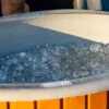 Wooden garden hot tub filled with ice cubes for cold plunge, recovery ice bath for athletes outdoors, ice in blue tub during sunset.
