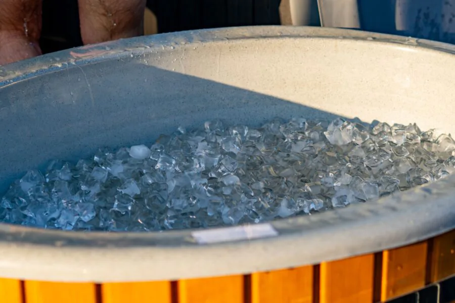 Wooden garden hot tub filled with ice cubes for cold plunge, recovery ice bath for athletes outdoors, ice in blue tub during sunset.