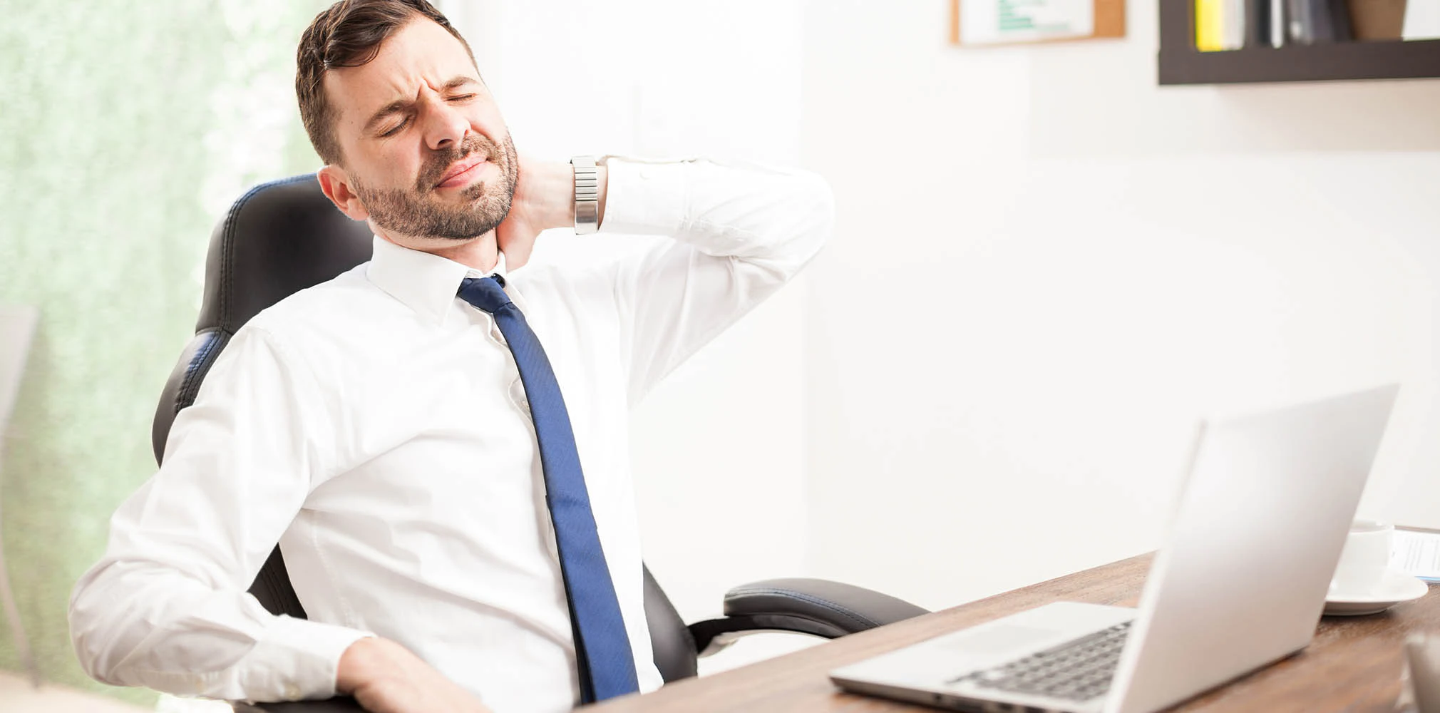A man grimaces in pain in his neck while working on his laptop.